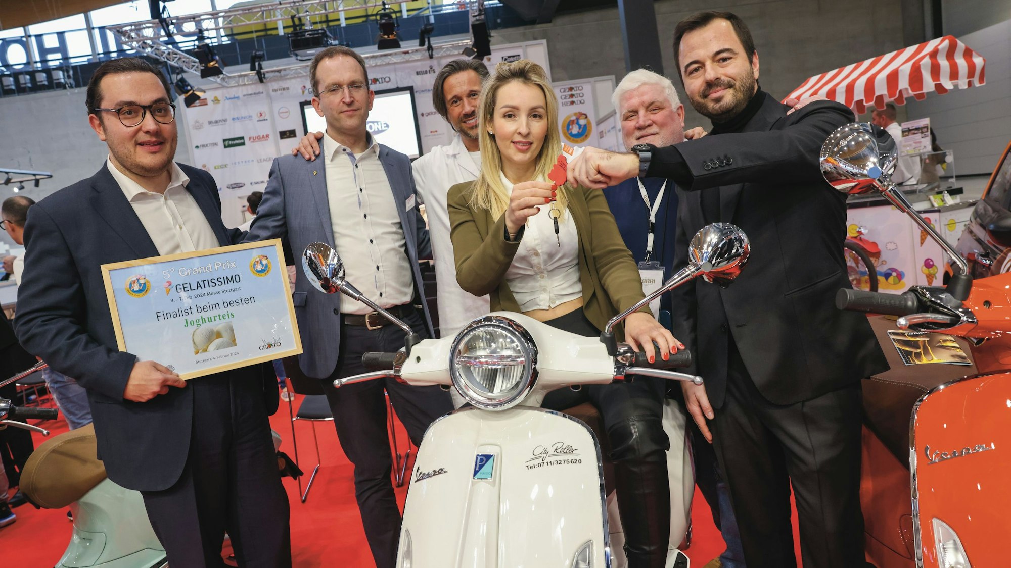 Angelika Szaban sitzt auf einer weißen Vespa. Um sie herum stehen fünf Männer, die mit ihr für ihre Auszeichnung posieren.