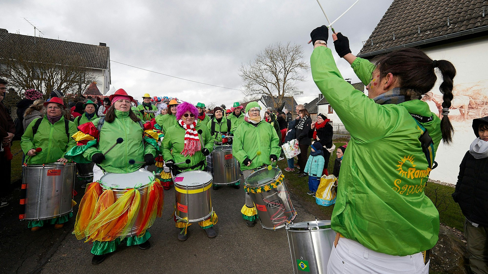 Eine Gruppe in grünen Jacken und bunten Kopfbedeckungen läuft mit großen Trommeln in einem Karnevalszug mit. Eine Frau mit langen dunklen Haaren gibt den Rhythmus vor.