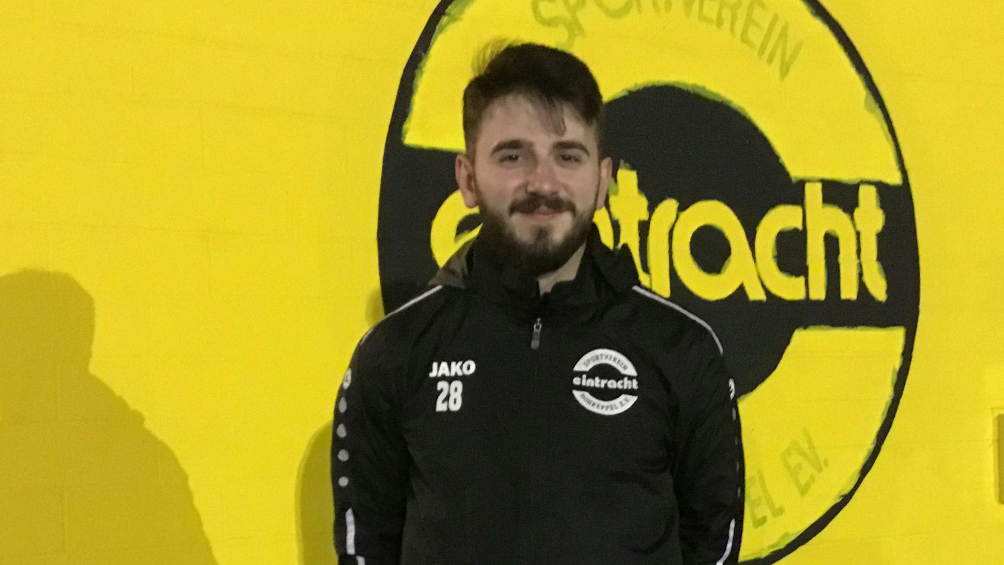 Zu sehen ist ein Fußballer vor einem großen Vereinswappen