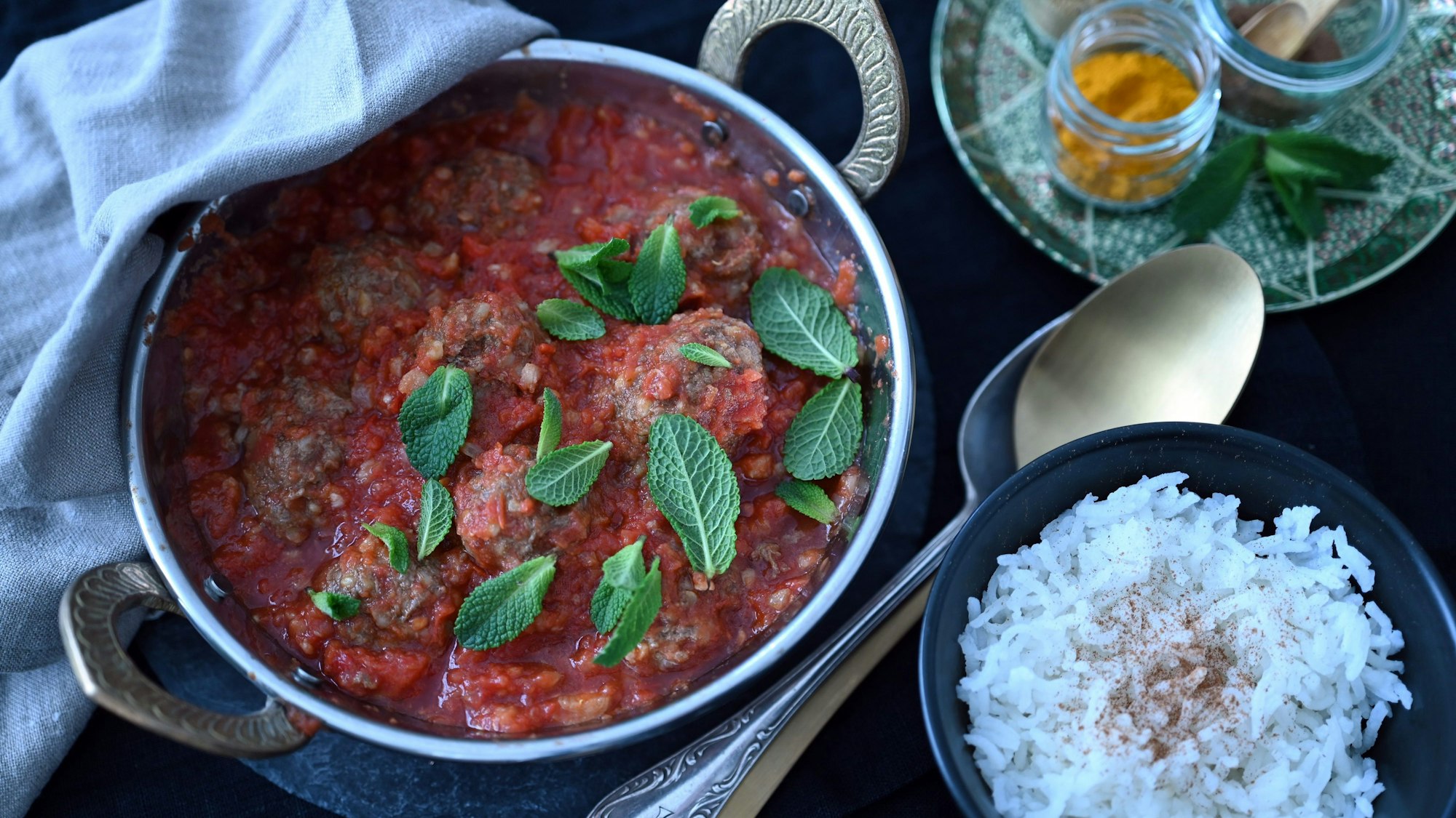 In einem Topf liegen Hackbällchen in einer Tomatensoße und mit Minzblättern überdeckt. In einer Schüssel ist Reis,