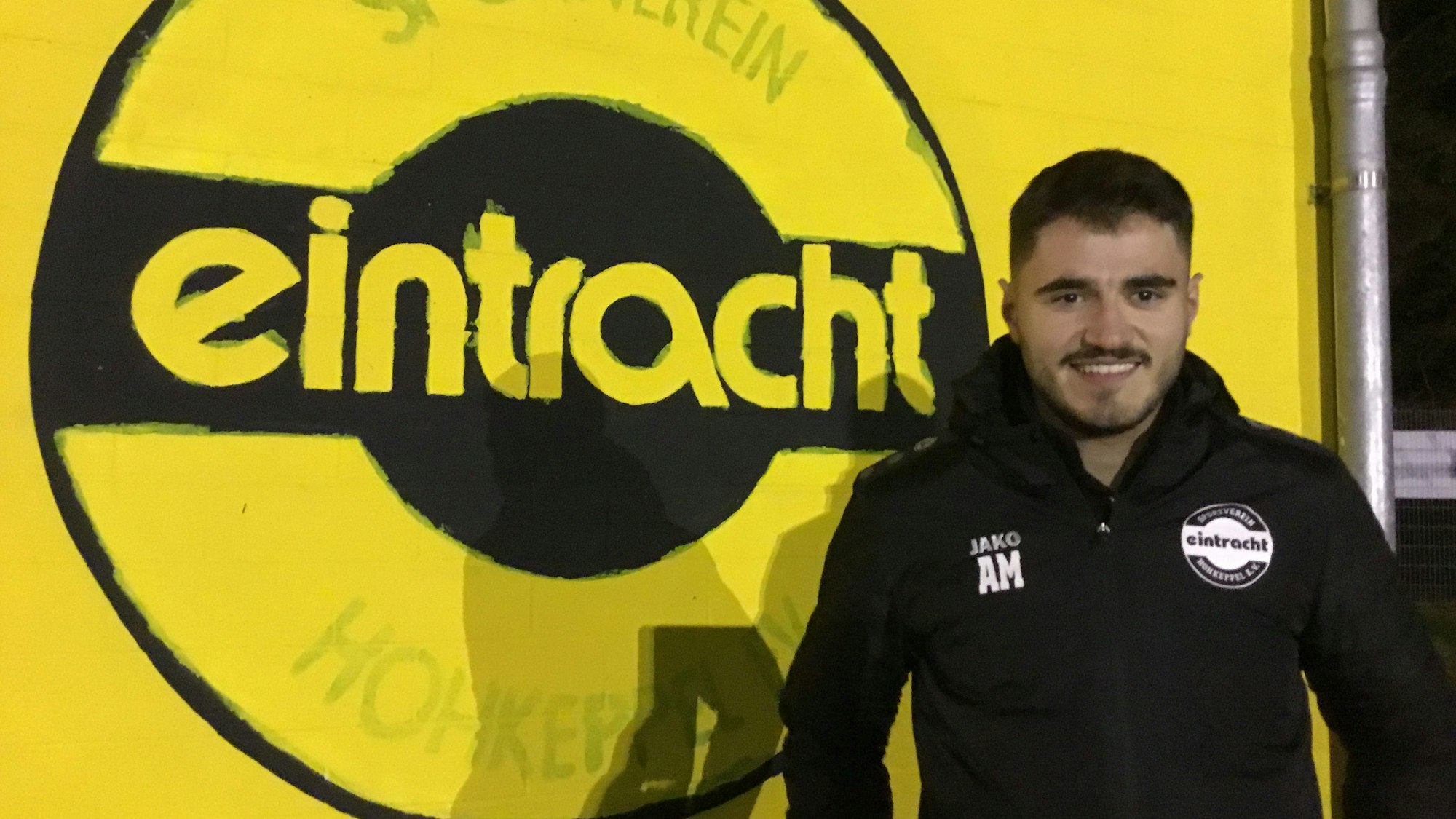 Zu sehen ist ein Fußballer vor einem großen Vereinswappen