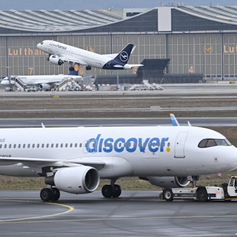 Während eine Maschine der Lufthansa startet, wird ein Passagierflugzeug der Discover Airlines am Frankfurter Flughafen von einem Schlepper gezogen.