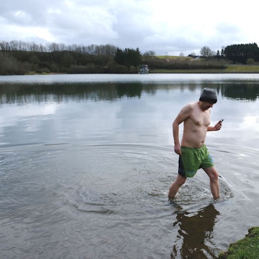 RRG-Redakteur Thorsten Wirtz mit Wollmütze und Badehose beim Verlassen des Freilinger Sees. In der Hand hält er ein Handy.