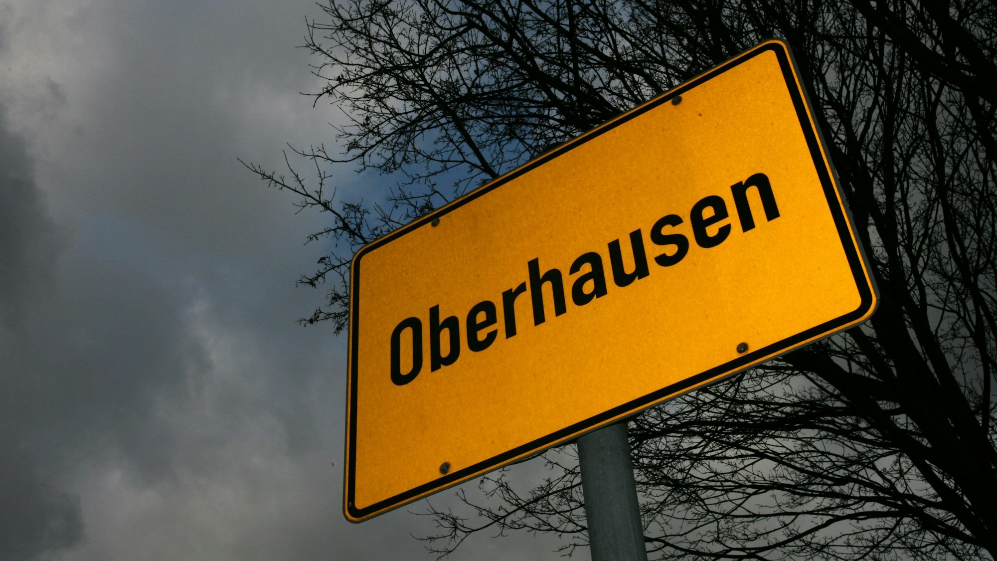 Das Ortsschild von Oberhausen steht vor düsterem Himmel und den dürren, blattlosen Ästen eines Baumes. (Symbolbild)