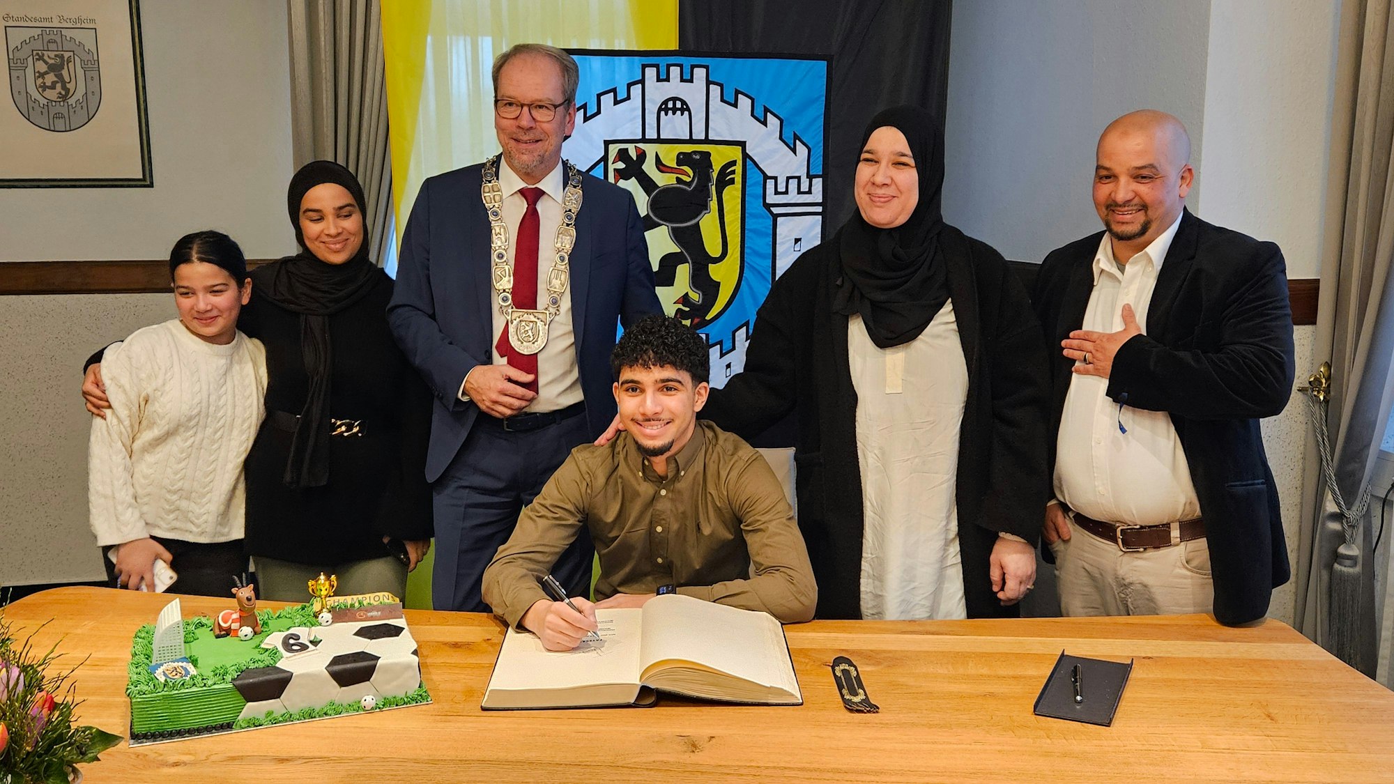 Ein junger Mann schreibt in ein großes Buch hinter ihm stehen seine Familie und ein Bürgermeister.