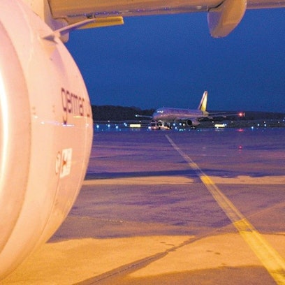 Das Bild zeigt die Turbine eines Flugzeuges und dahinter Vorfeld sowie Start- und Landebahn mit einem weiteren Flugzeug.