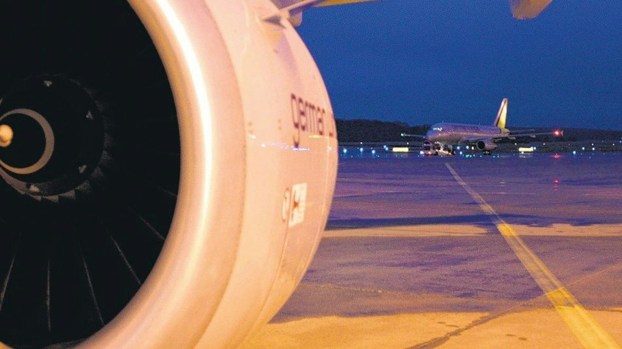 Das Bild zeigt die Turbine eines Flugzeuges und dahinter Vorfeld sowie Start- und Landebahn mit einem weiteren Flugzeug.