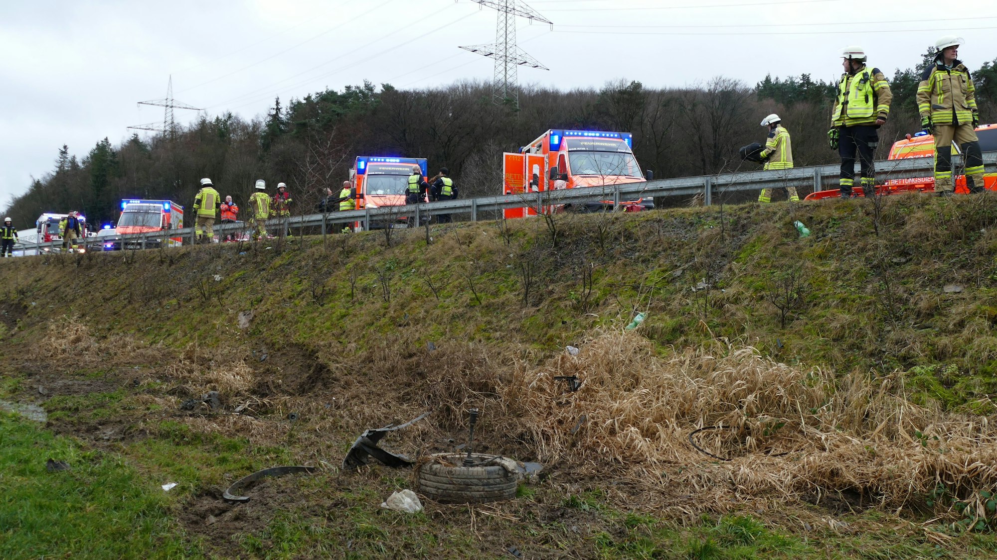 Trümmerteile eines Fahrzeugs liegen auf einer Wiese. An einem steilen Hang stehen viele Rettungsfahrzeuge und Feuerwehrleute.