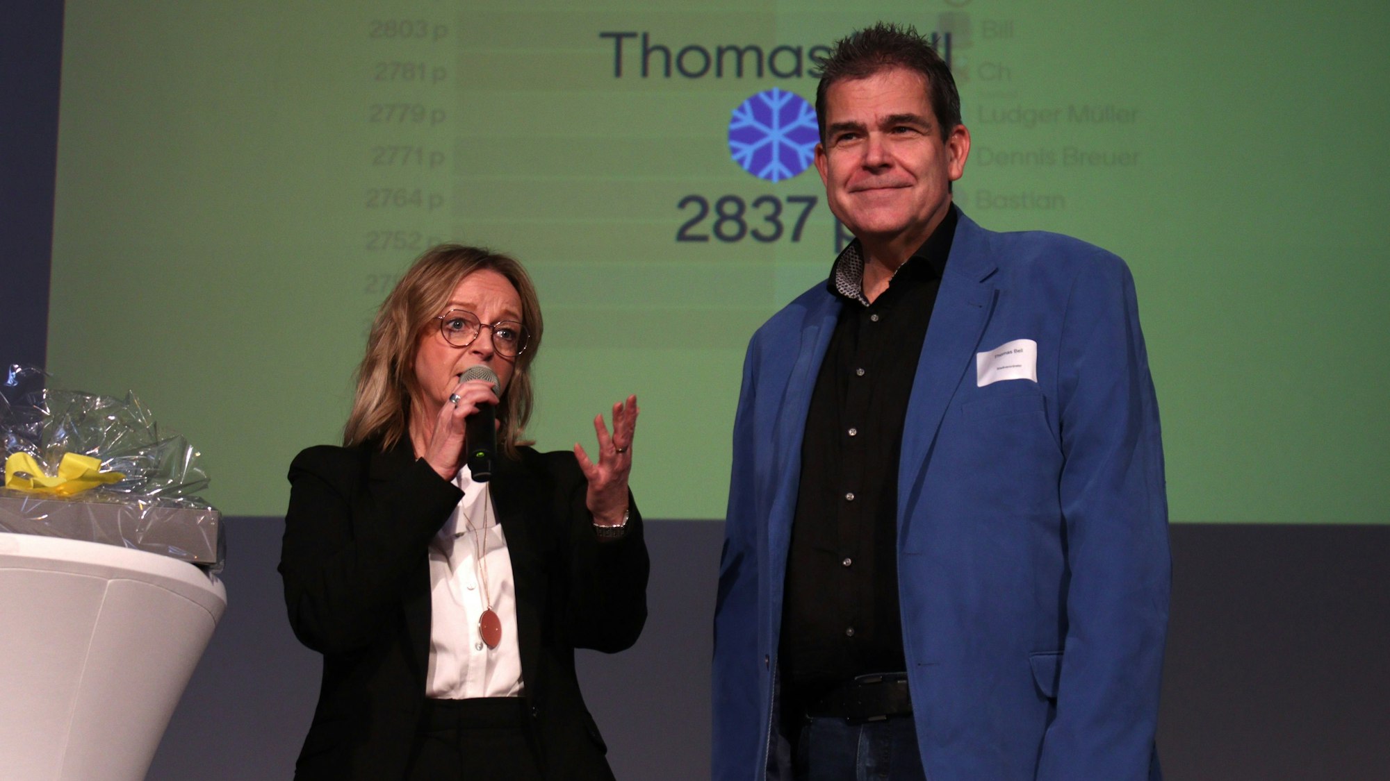 Thomas Bell steht mit Sabine Preiser-Marian, die ein Mikrofon in der Hand hält, auf der Bühne.