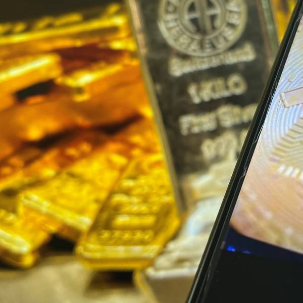 Auf einem Bildschirm sind die Edelmetalle Gold und Silber zu sehen, davor ist ein Smartphone-Bildschirm zu sehen, auf dem das Logo der Kryptowährung Bitcoin zu sehen ist.