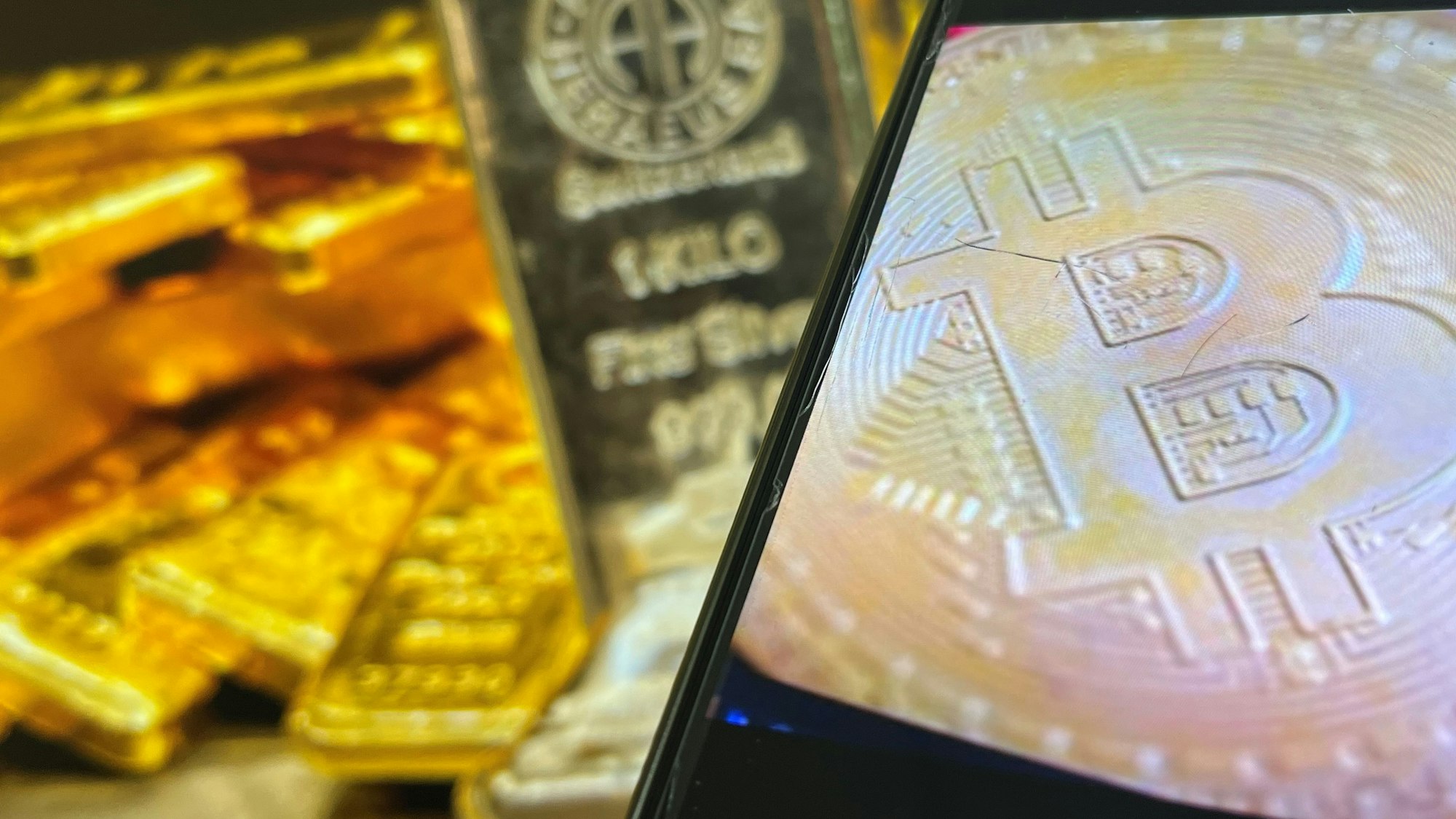 Auf einem Bildschirm sind die Edelmetalle Gold und Silber zu sehen, davor ist ein Smartphone-Bildschirm zu sehen, auf dem das Logo der Kryptowährung Bitcoin zu sehen ist.