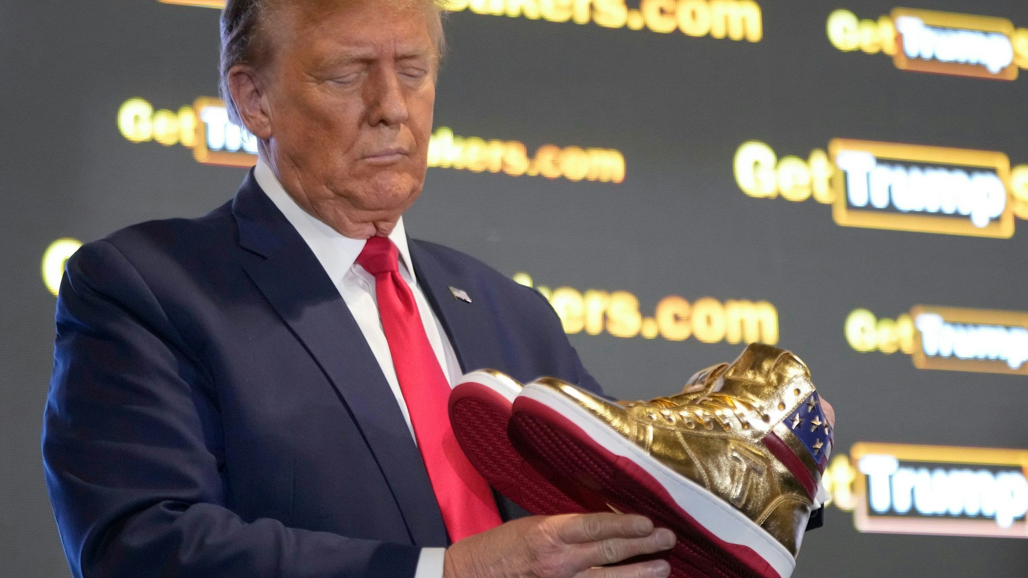 Donald Trump hält goldene Trump-Sneakers in Händen.