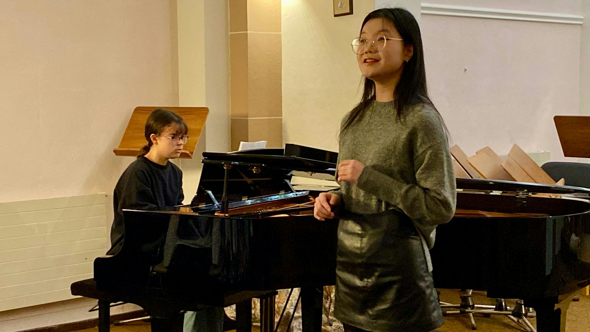 Zu sehen sind zwei junge Frau, eine sitzt an einem Flügel, die andere steht daneben, sie singt.