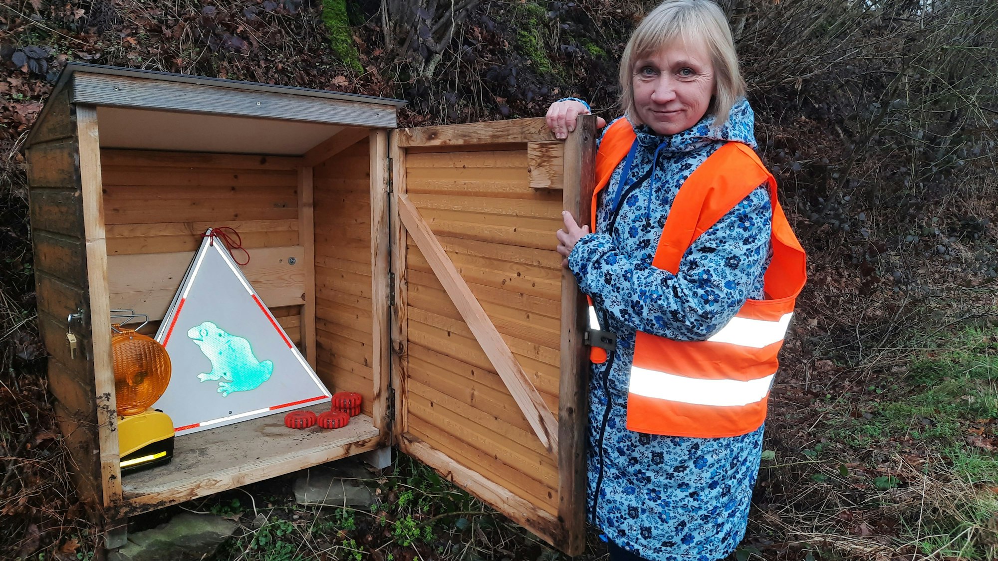 Eine Frau mit Warnweste hält einen Holzschrank geöffnet, darin sind Warnschilder und Leuchten.