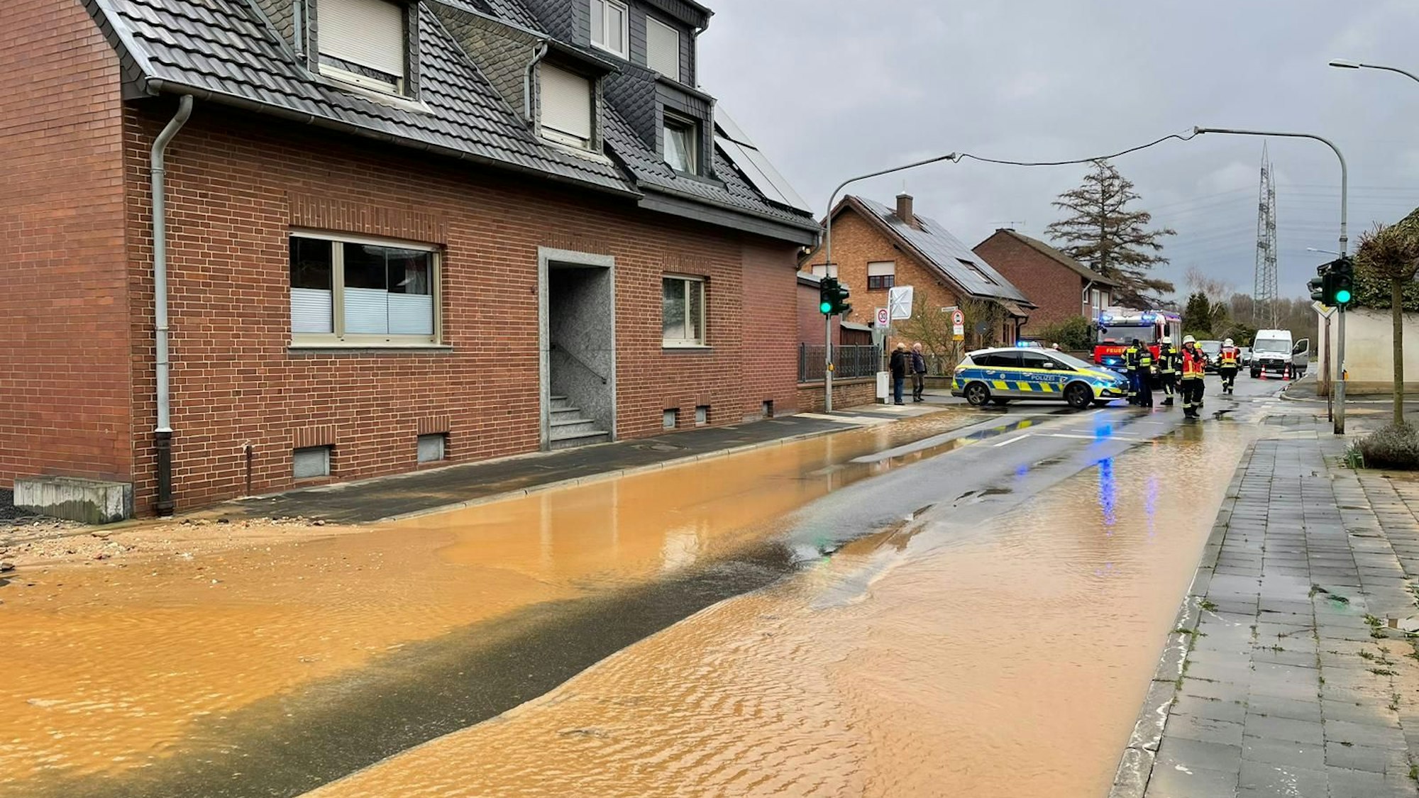 Auf einer Straße steht schmutzig-braunes Wasser, im Hintergrund sind ein Polizei- und ein Feuerwehrfahrzeug zu sehen.