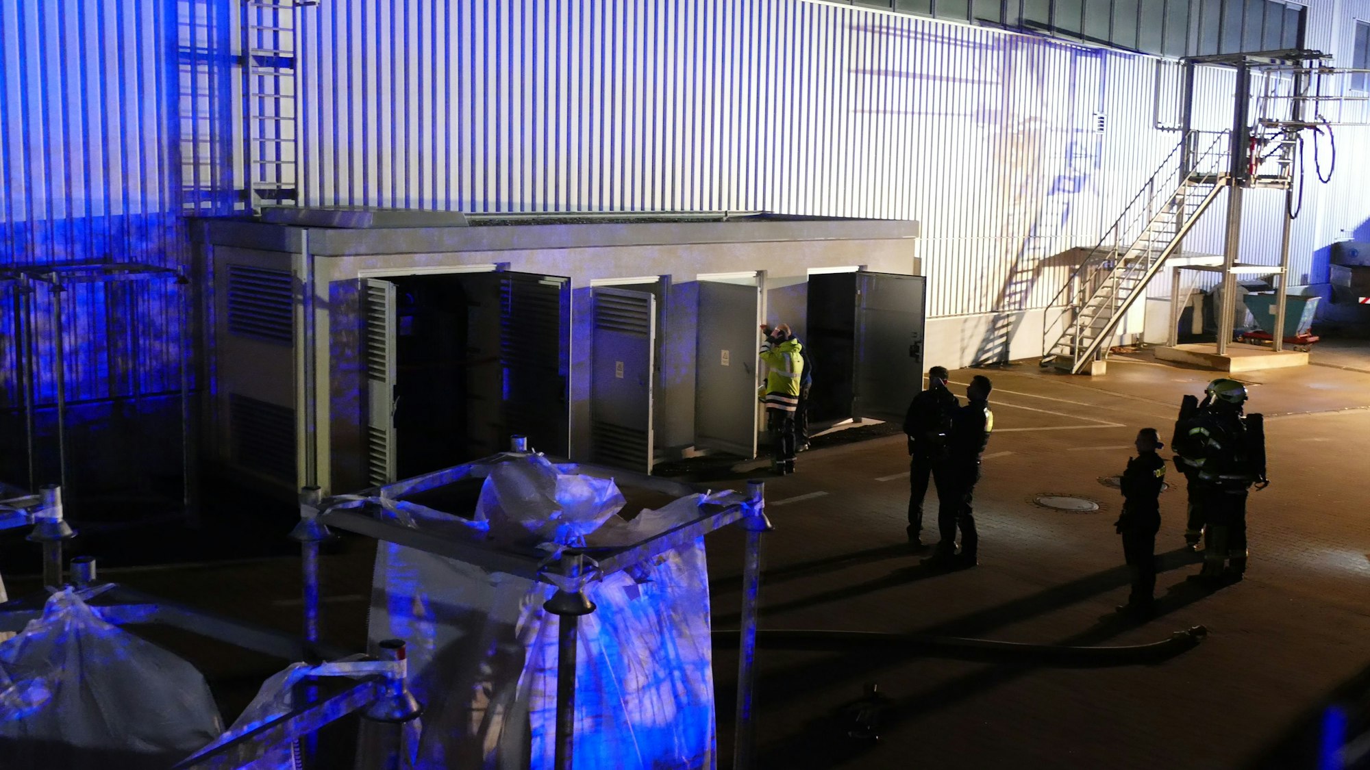 Feuerwehrleute stehen vor einem Trafo-Häuschen und einer riesigen Halle.