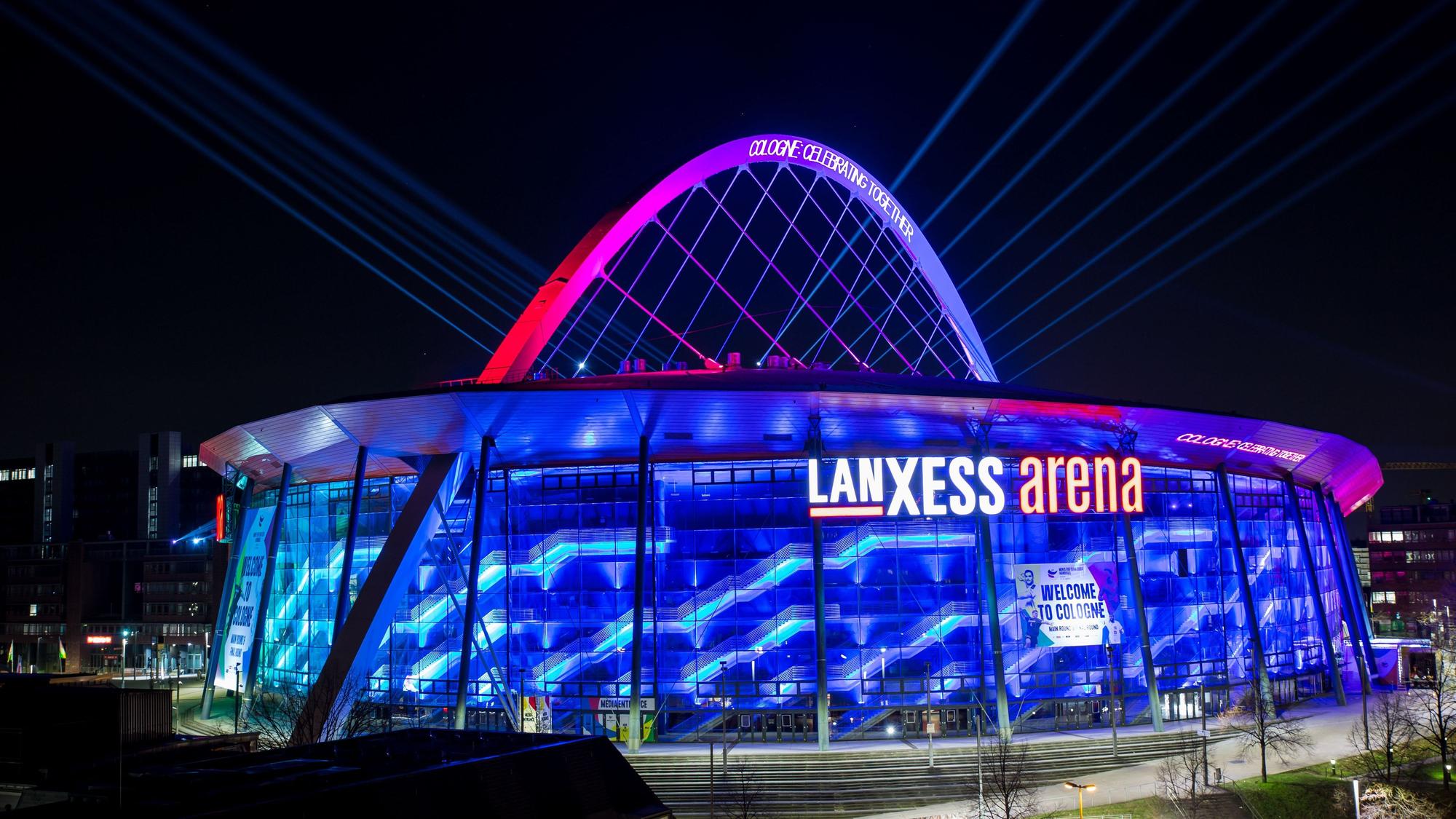 Illumination der Lanxess-Arena zur Handball-Europameisterschaft im Januar. Die Halle erstrahlt in blauem Licht, der Bogen ist rosa angestrahlt.