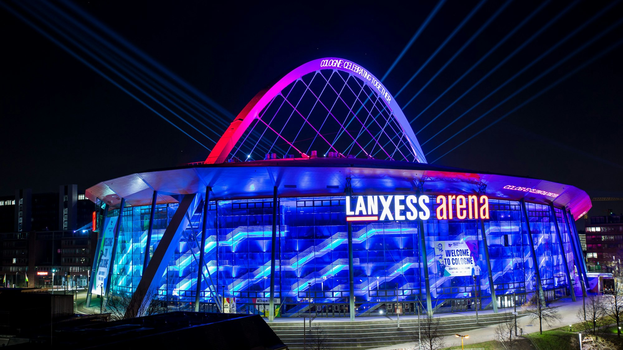 Illumination der Lanxess-Arena zur Handball-Europameisterschaft im Januar. Die Halle erstrahlt in blauem Licht, der Bogen ist rosa angestrahlt.