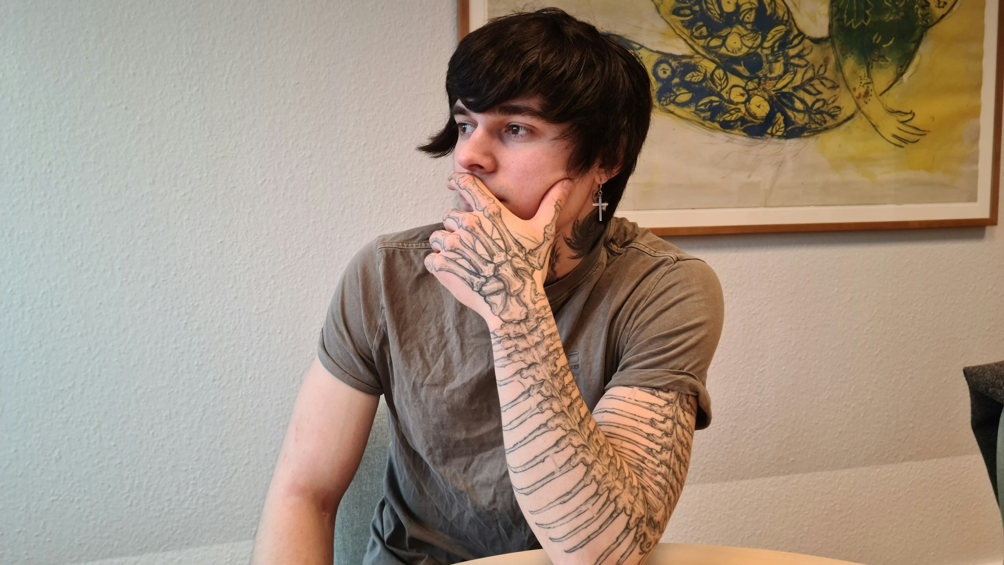 Ein junger Mann sitzt an einem Tisch. Sein aufgestützter Arm ist von oben bis unten tätowiert.