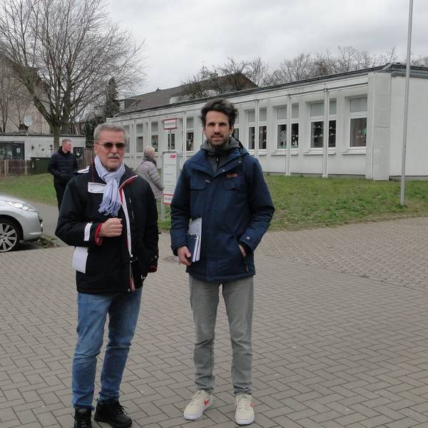 Ein älterer und ein jüngerer Mann stehen vor einem Schulgrundstück.