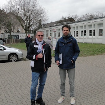 Ein älterer und ein jüngerer Mann stehen vor einem Schulgrundstück.