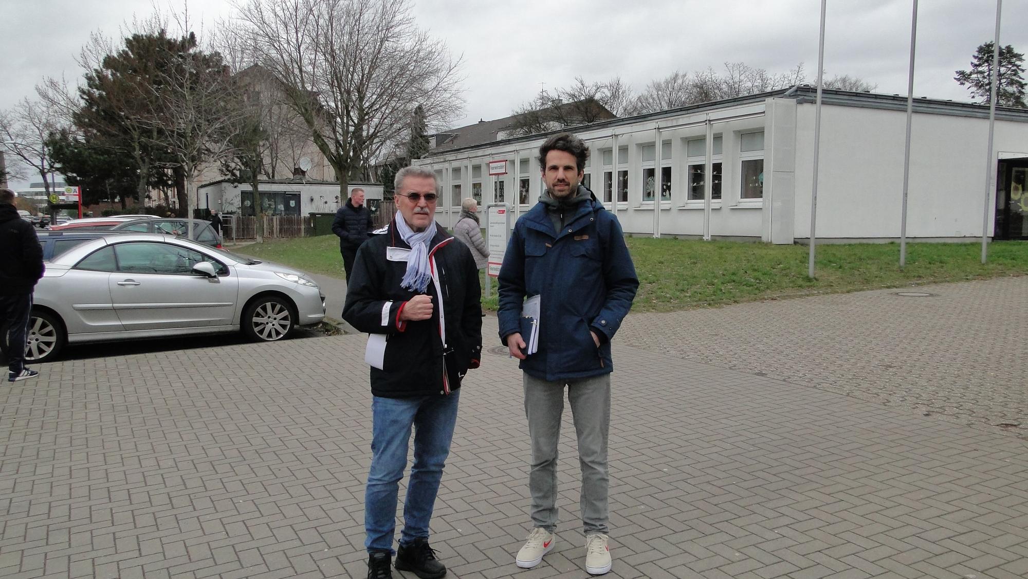 Ein älterer und ein jüngerer Mann stehen vor einem Schulgrundstück.