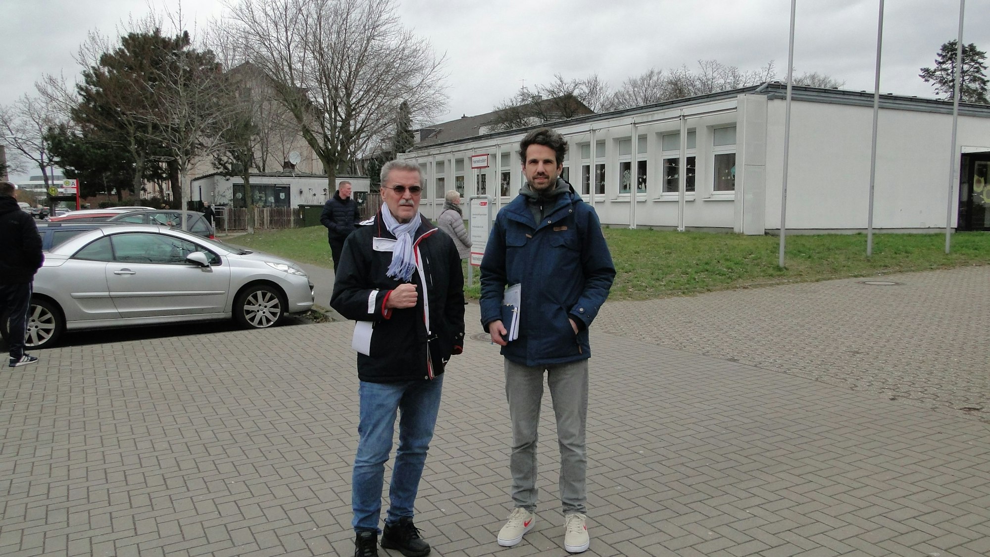 Ein älterer und ein jüngerer Mann stehen vor einem Schulgrundstück.