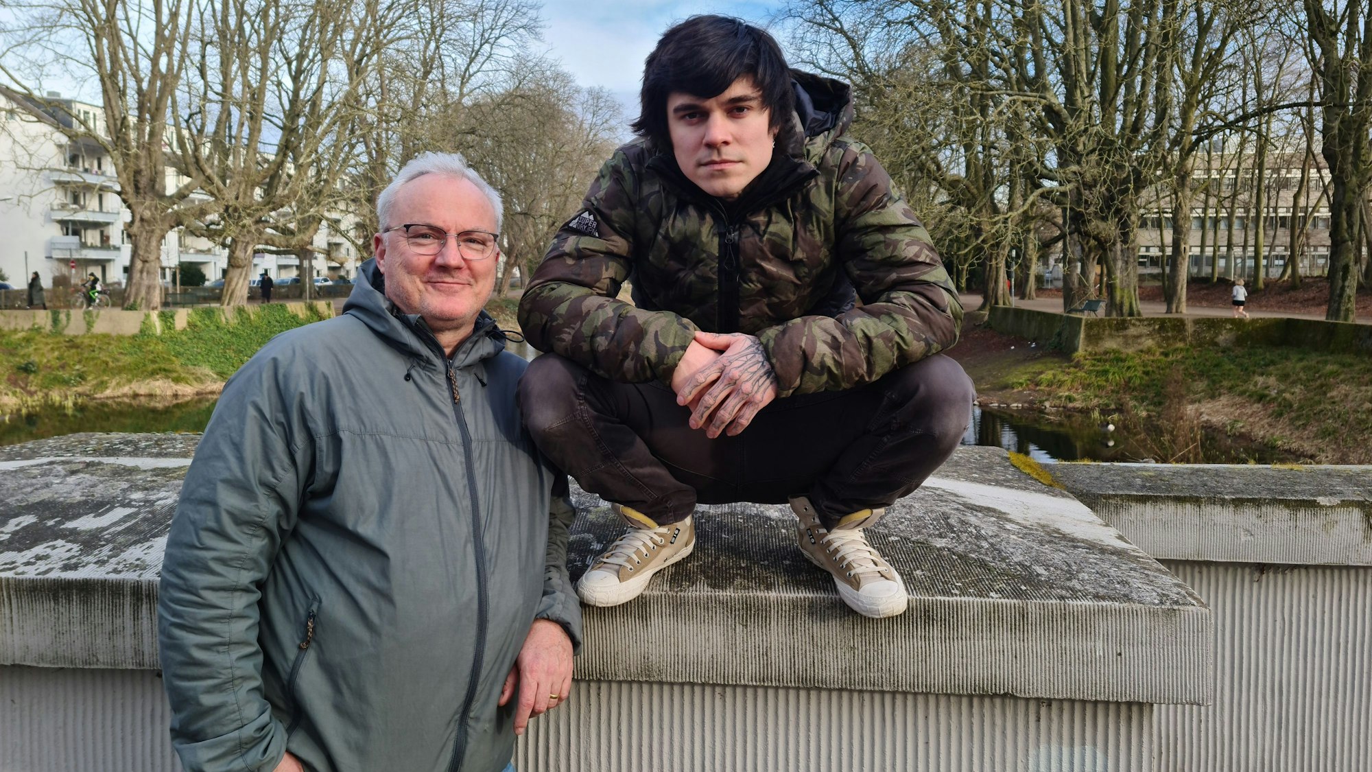 Ein Mann mit grauem Haar steht neben auf einer Mauer hockenden jüngeren Mann.