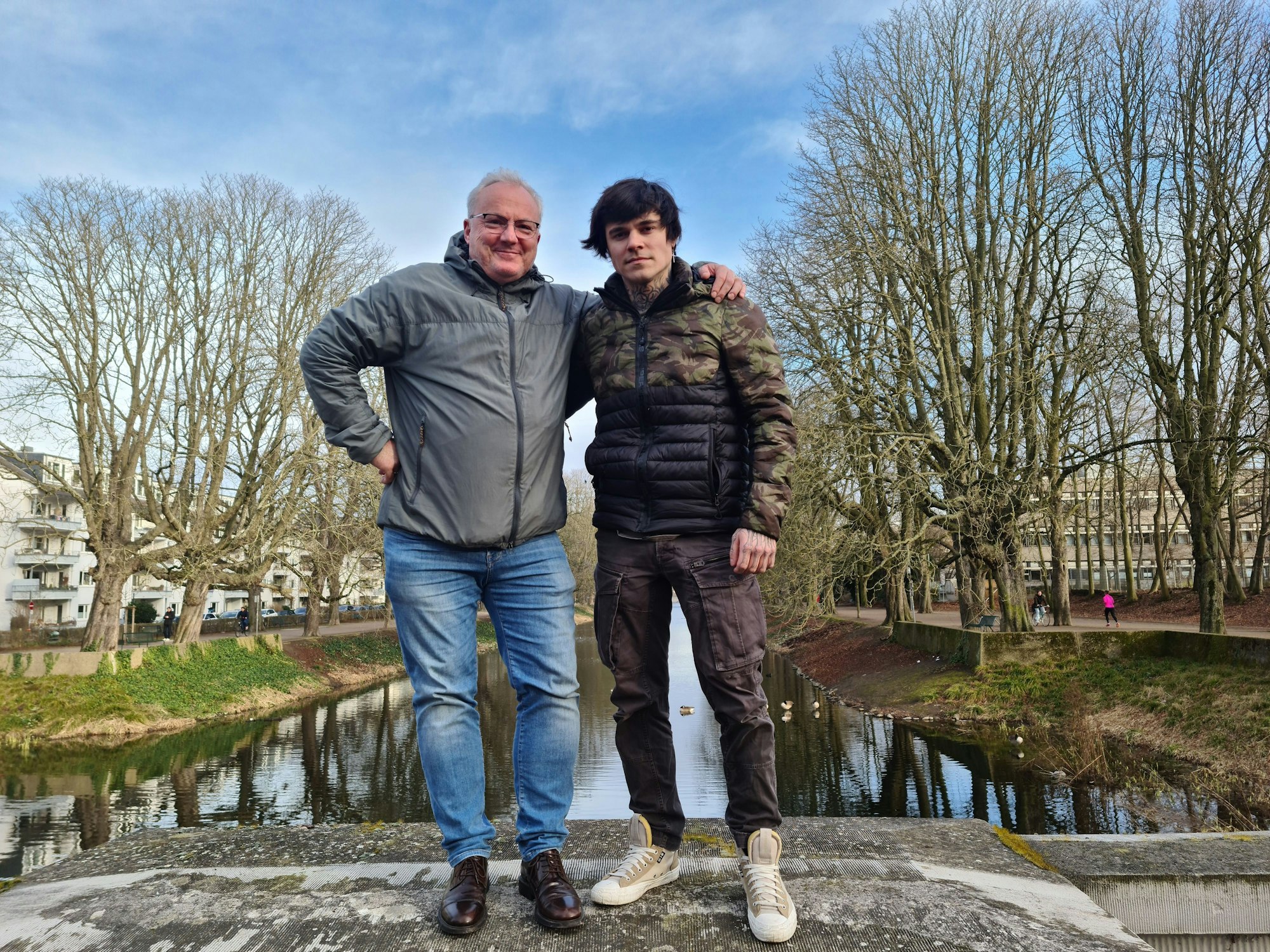 Ein älterer und ein jüngerer Mann stehen nebeneinander auf einer Mauer über dem Lindenthaler Kanal.