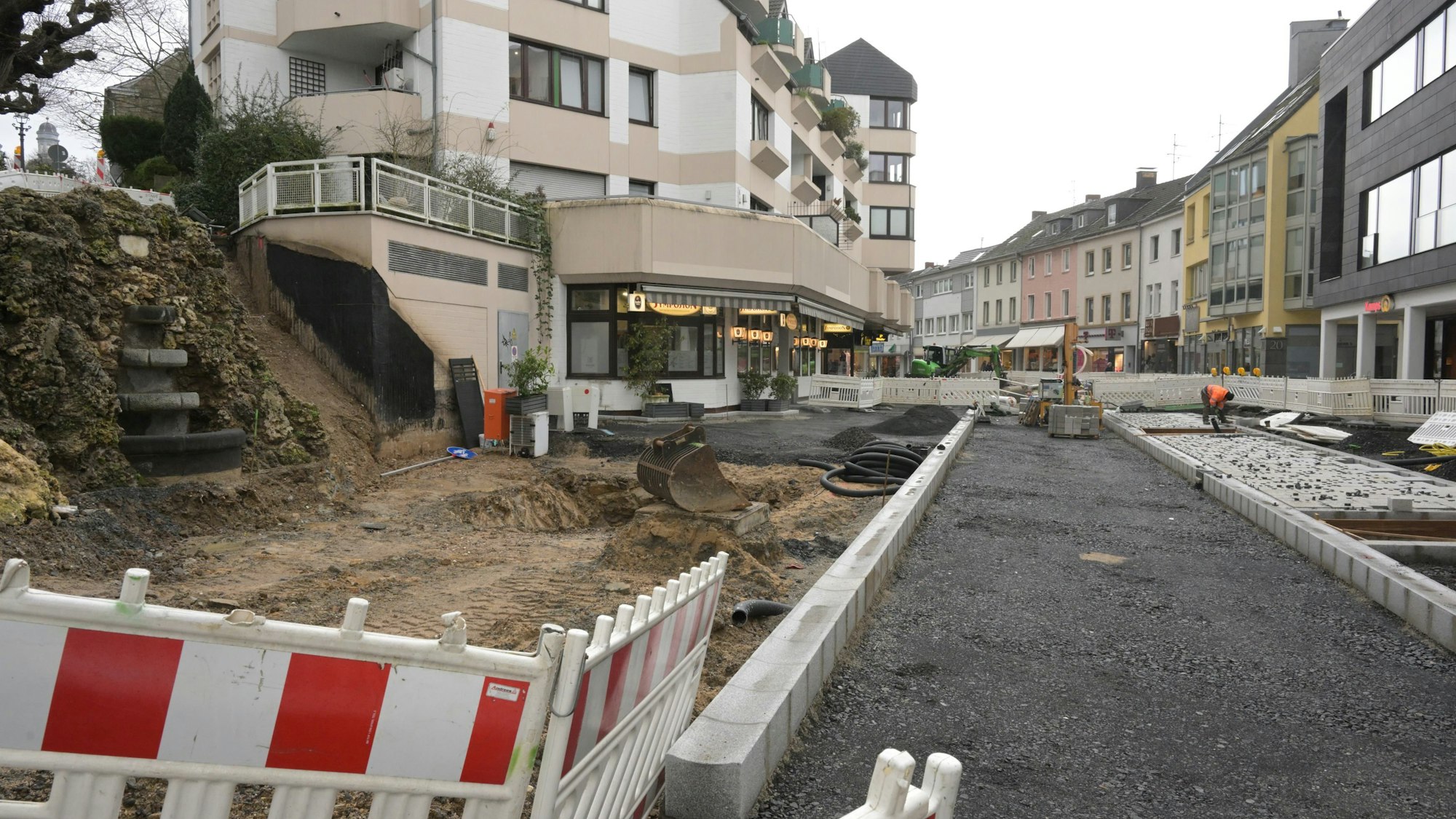 Die Fahrbahn ist bereits mit Asphalt ausgekoffert, rechts und links sind Randsteine verlegt. Ein Teil des Fußwegs rechts ist schon mit dem neuen Pflaster ausgestattet. Links vor dem Emilienbrunnen ist noch Erde. Im Vordergrund steht eine rot-weiße Bake.