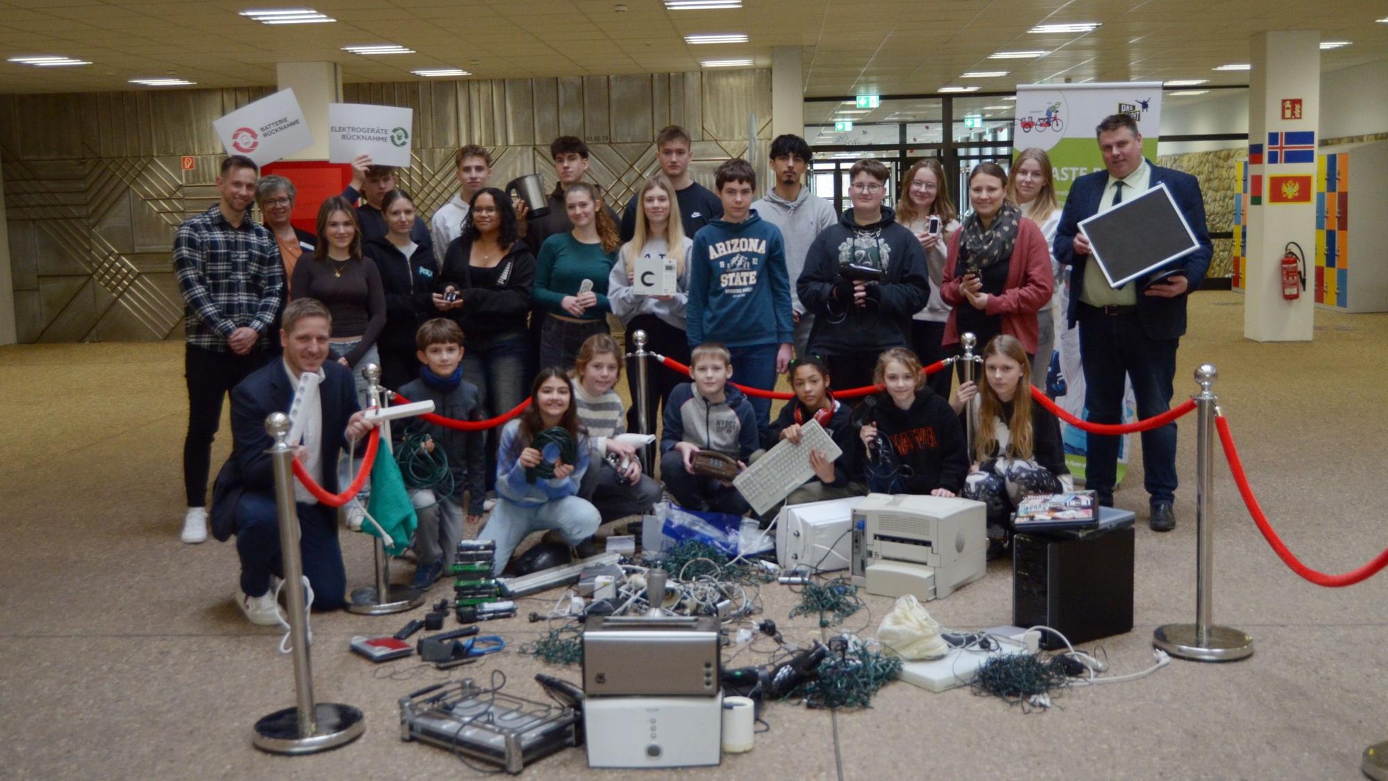 Schülerinnen, Schüler, Lehrer, Lehrerinnen und Landrat Markus Ramers haben sich zu einem Gruppenbild formiert, vor ihnen ausrangierte Elektrogeräte wie Drucker und Toaster sowie Kabel und Lichterketten.
