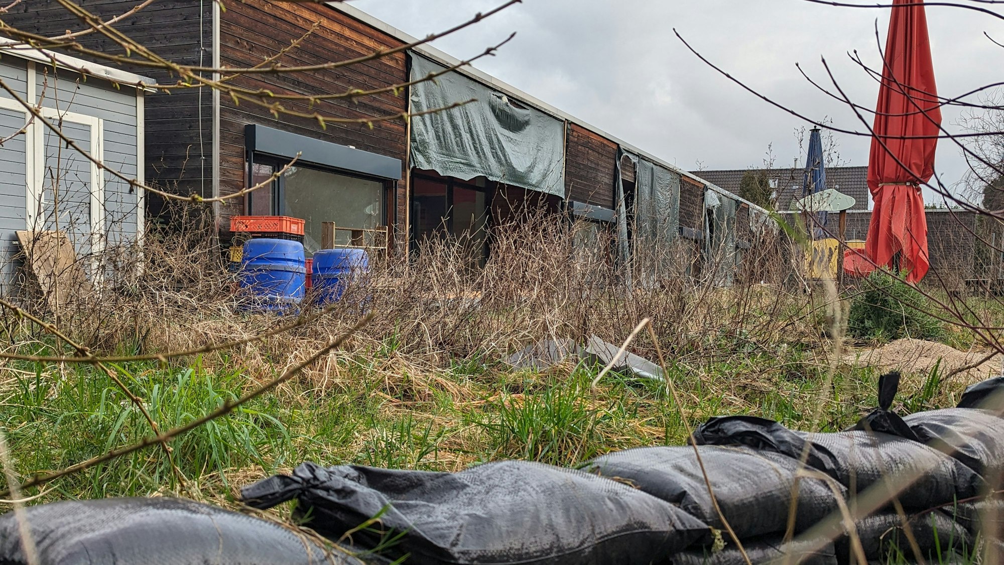 Ein verwilderter Außenbereich vor der zum Teil mit Plastikplanen verhängten Holzfassade einer Kita, im Vordergrund Sandsäcke.