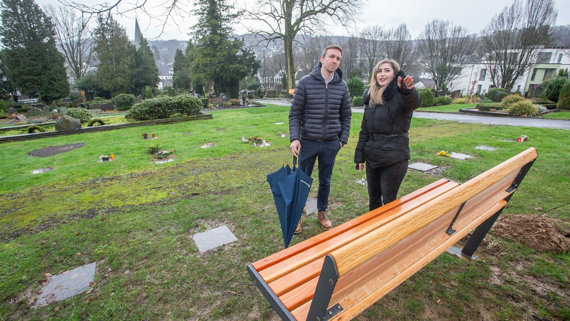 Ein Mann und eine Frau stehen vor einer Bank auf dem Friedhof.