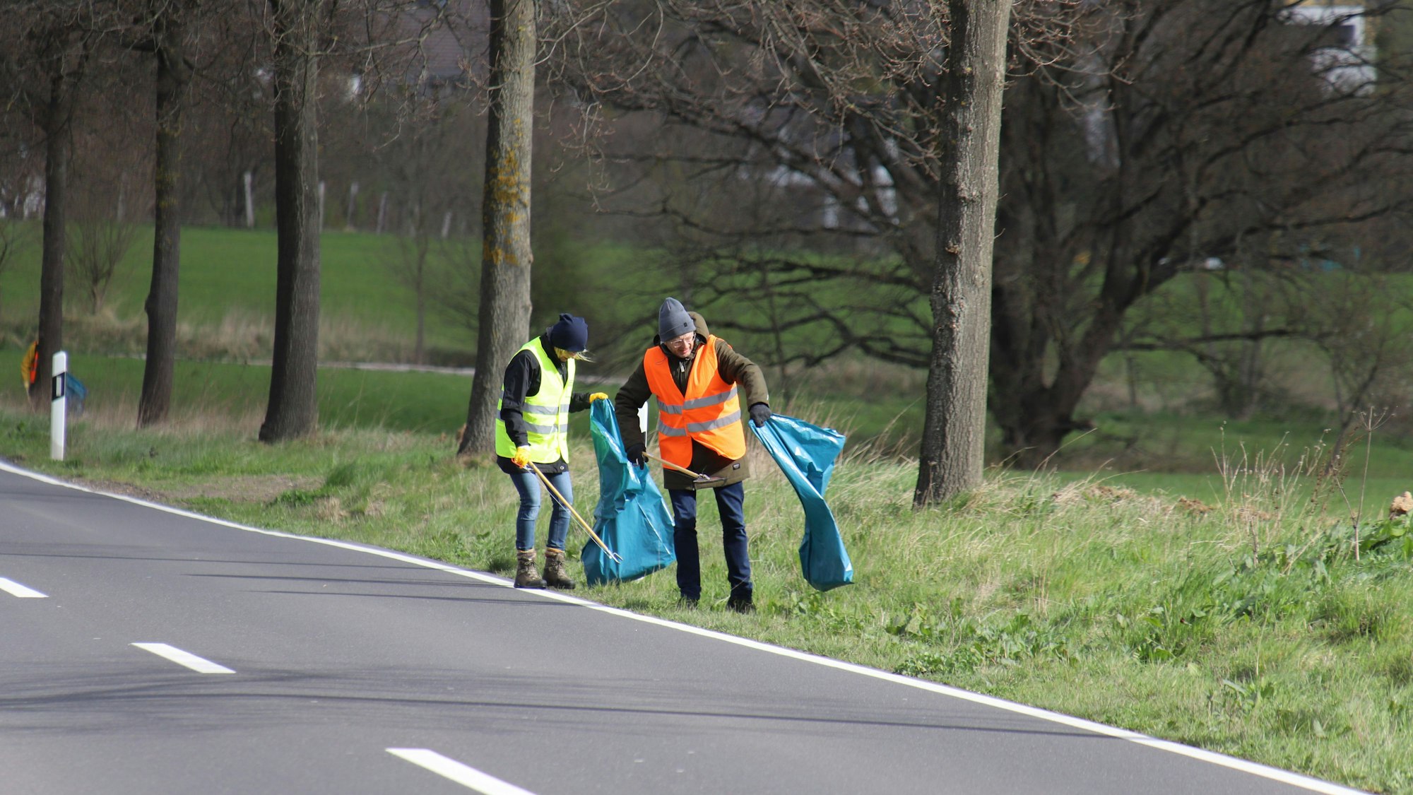 Zwei Menschen in Warnwesten und mit langen Zangen und blauen Müllsäcken gehen an einem Straßenrand und sammeln Müll.