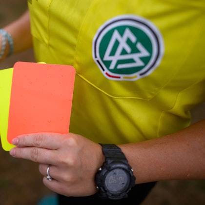 Eine Schiedsrichterin, steht mit roter und gelber Karte auf einem Rasen und hält in der anderen Hand eine Trillerpfeife.