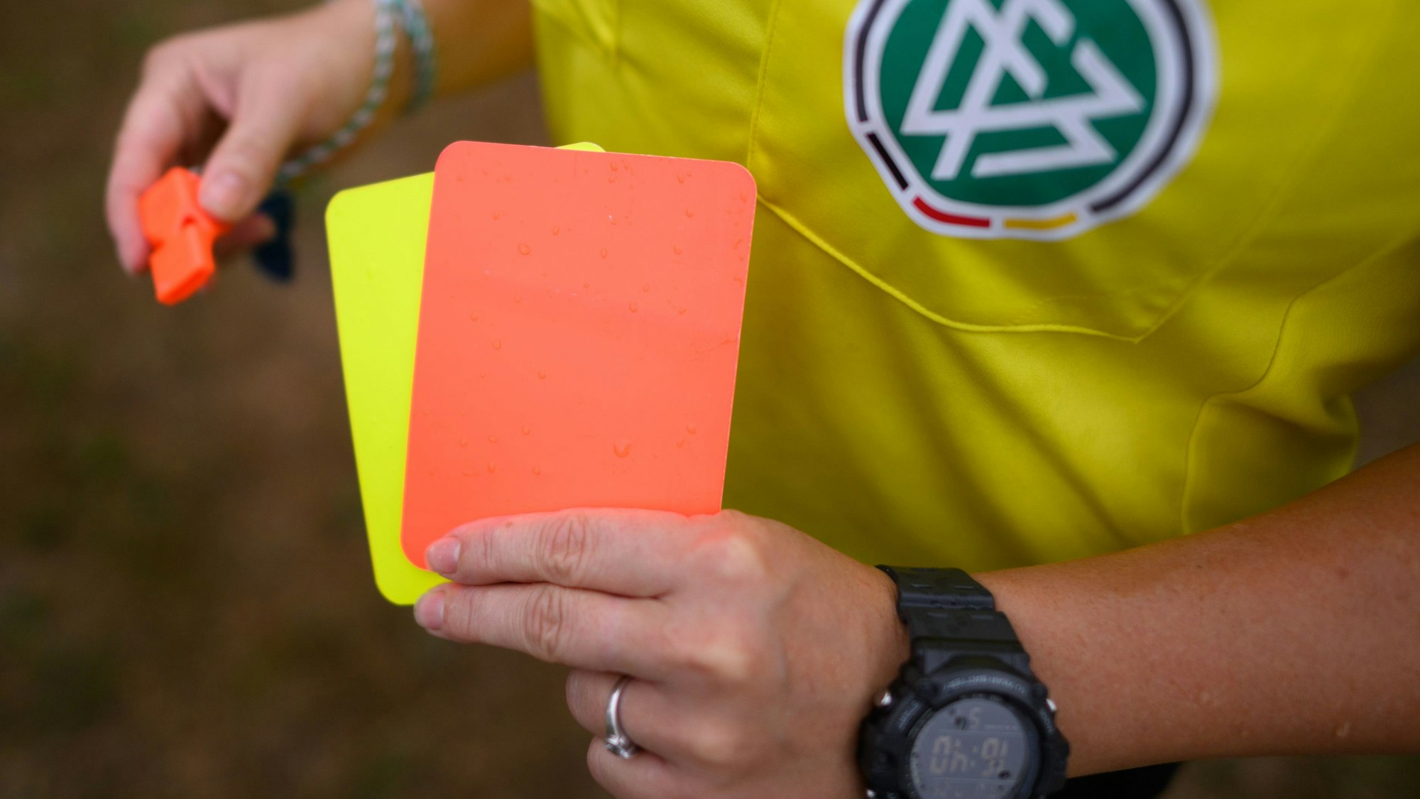 Eine Schiedsrichterin, steht mit roter und gelber Karte auf einem Rasen und hält in der anderen Hand eine Trillerpfeife.