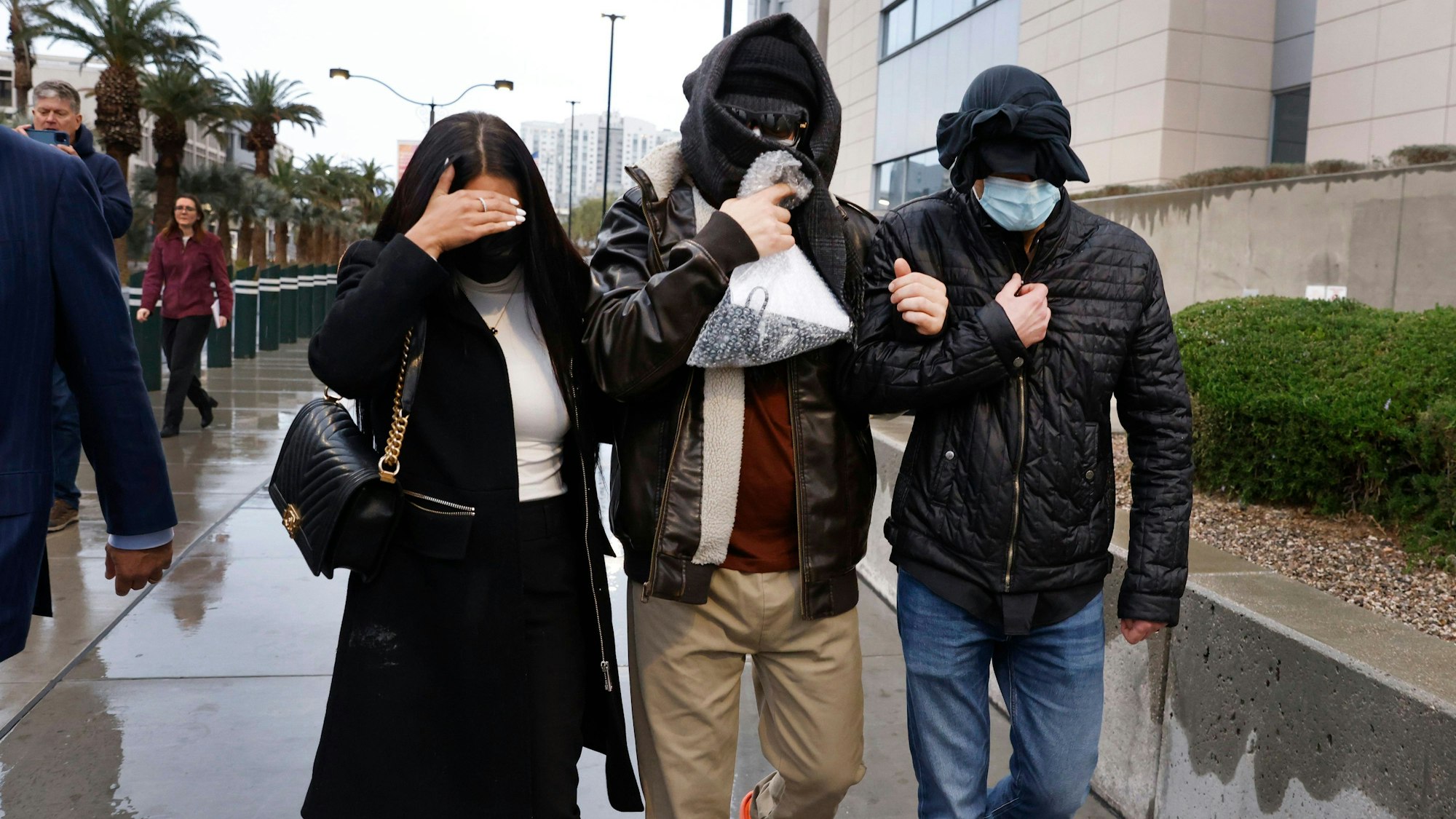 Drei eingehakte Personen gehen maskiert über eine verregnete Straße - im Hintergrund stehen Palmen und Gebäude.