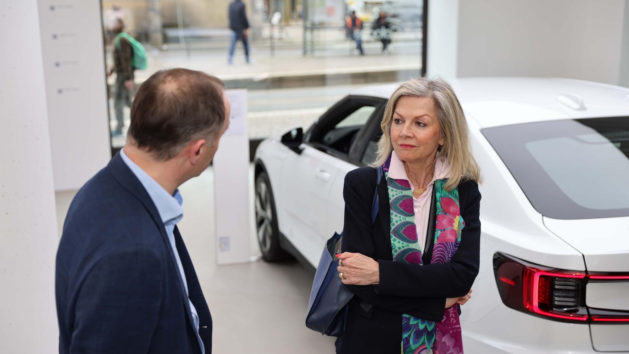 Eine blonde Frau im Gespräch mit einem Mann vor einem weißen Auto.