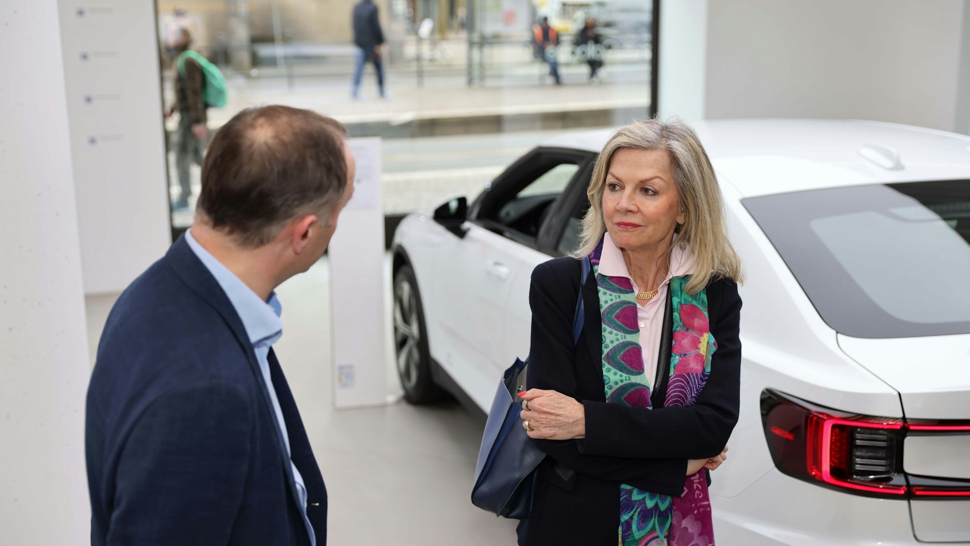 Eine blonde Frau im Gespräch mit einem Mann vor einem weißen Auto.