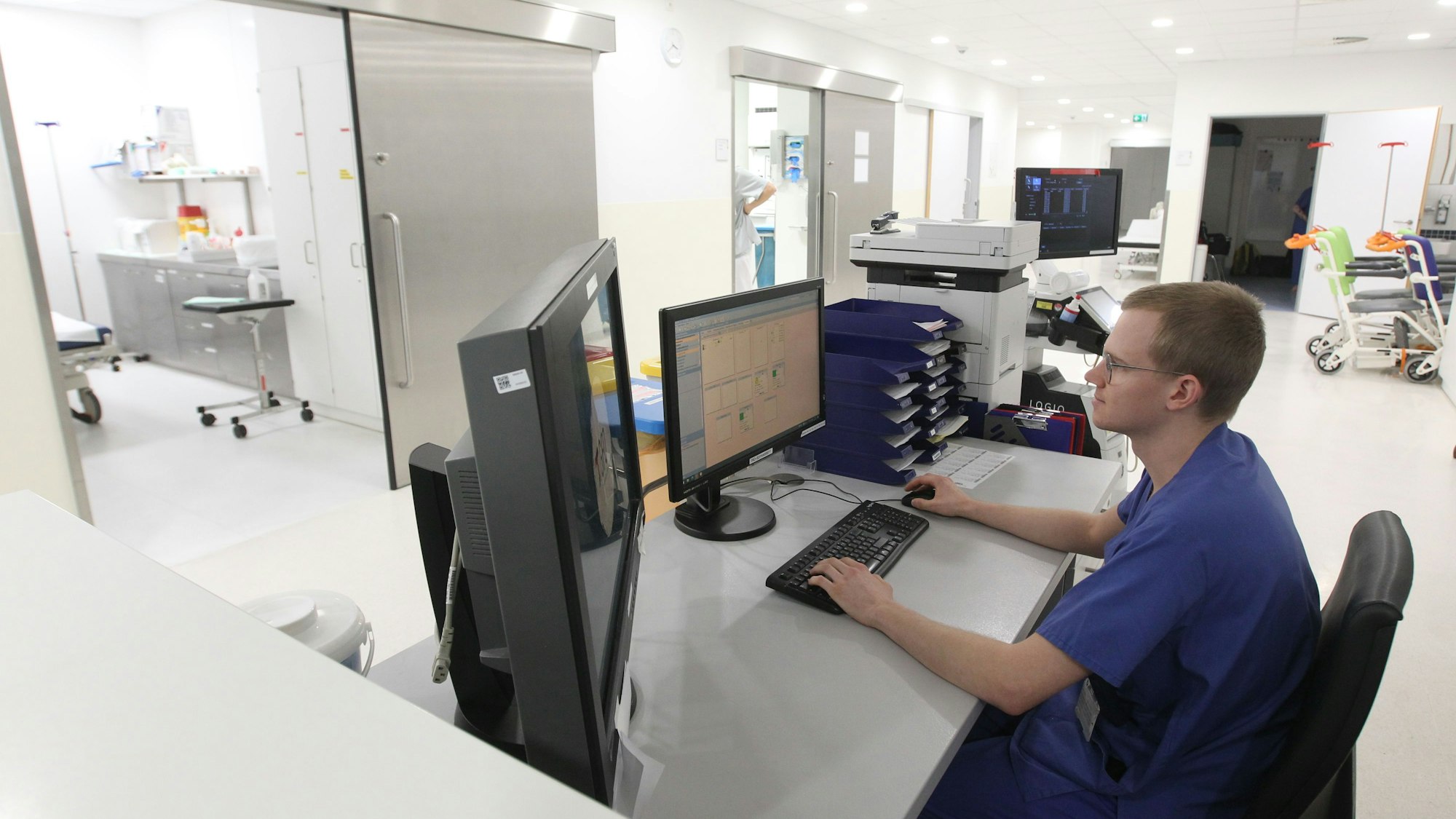 Ein junger Mann in blauer Kleidung eines Pflegers sitzt an einem Schreibtisch mit zwei Bildschirmen. Im Hintergrund sind Räume mit medizinischem Gerät zu sehen.