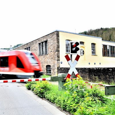 Ein Zug der Regionalbahn 25 passiert den Bahnübergang Märkische Straße in Engelskirchen, im Hintergrund die Fabrik Ermen und Engels, wo heute das Industriemuseum und das Rathaus zu finden sind.