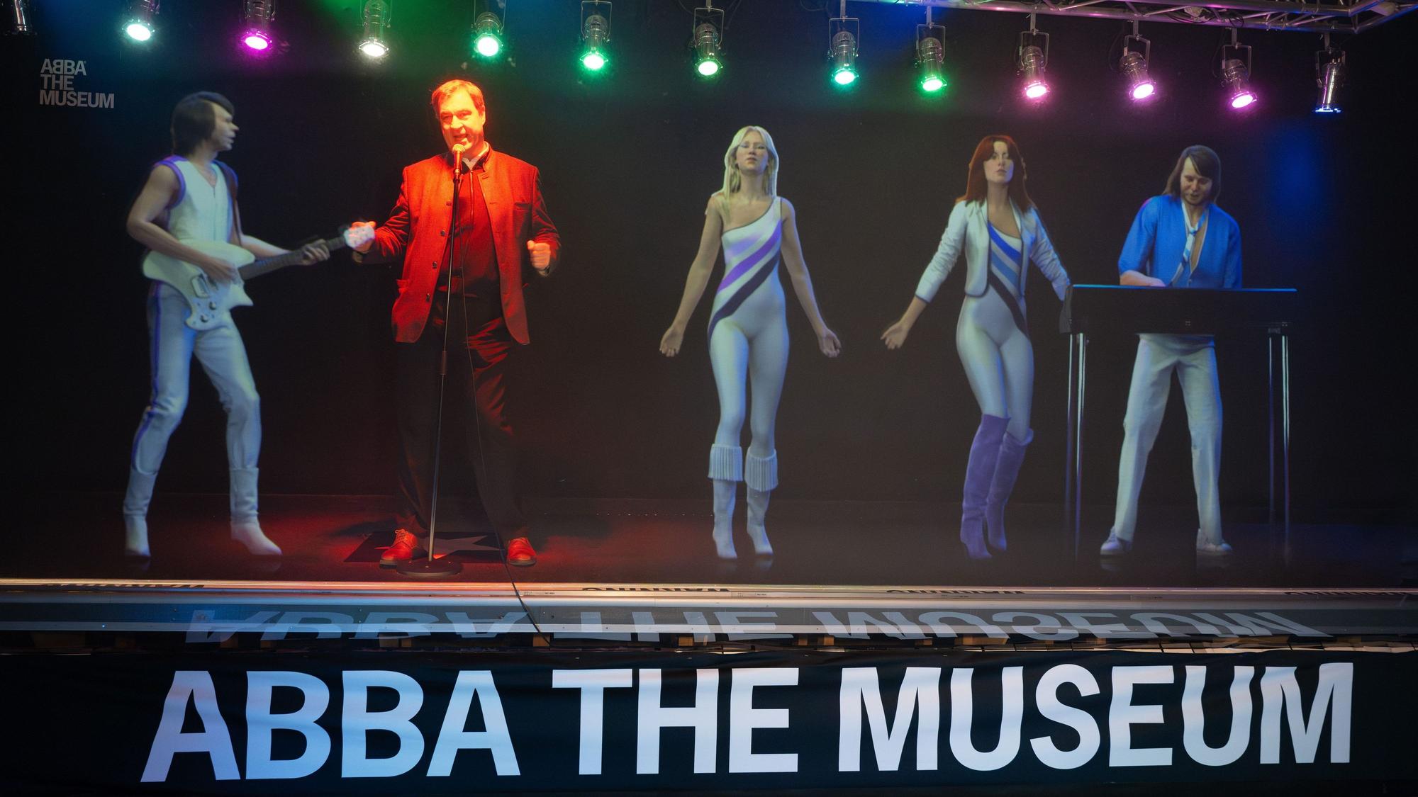 Bayerns Ministerpräsident Söder steht auf einer Bühne. Er singt und tanzt mit ABBA-Hologrammen.