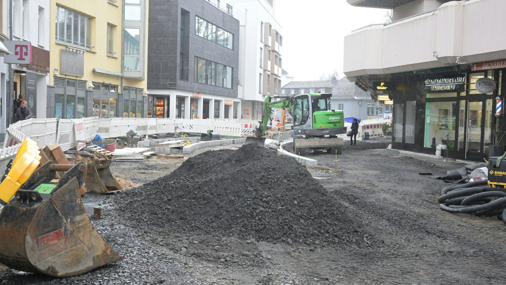 Im Vordergrund sieht man eine Baggerschaufel und einen Berg von schwarzem Schotter. Der Gehweg links ist durch Absperrgitter abgesperrt.