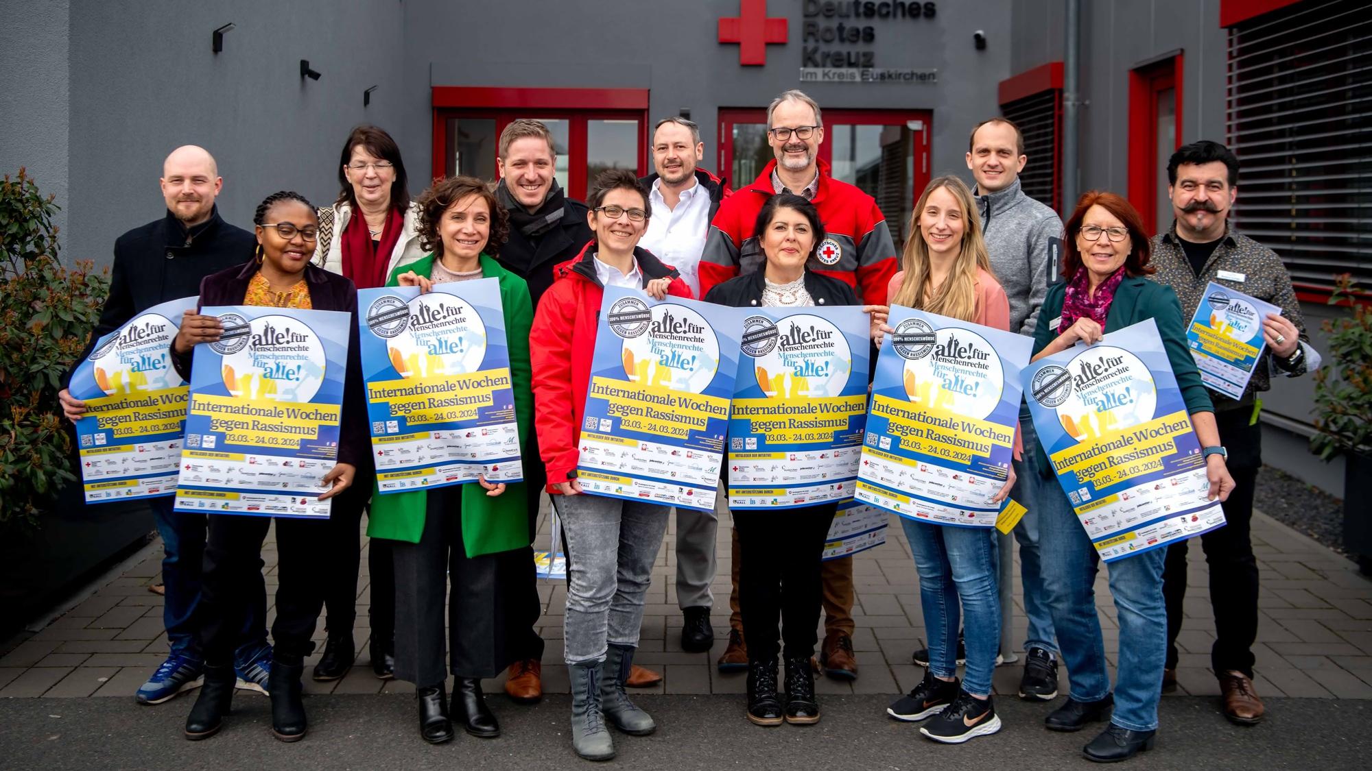 Eine Gruppe Männer und Frauen steht vor dem DRK-Zentrum in Euskirchen. Einige halten Plakate in den Händen, die auf die Aktionswochen gegen Rassismus hinweisen.