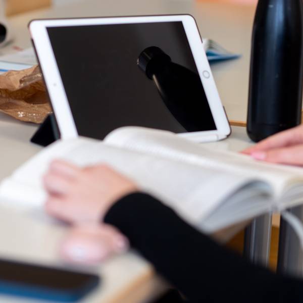 Das Bild zeigt eine Schülerin, die in einem Buch liest. Hinter dem Buch steht ein Tablet.