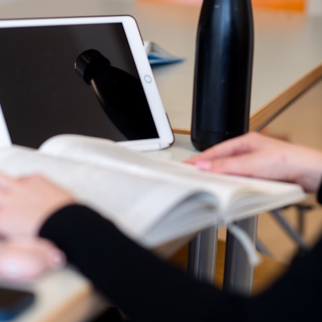 Das Bild zeigt eine Schülerin, die in einem Buch liest. Hinter dem Buch steht ein Tablet.