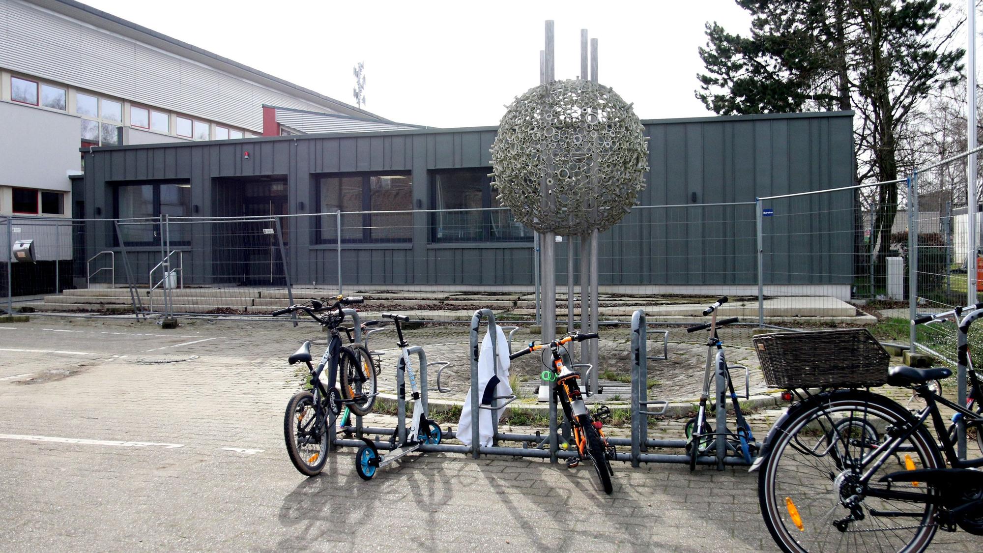 Im Vordergrund ist eine Metallskulptur in Form einer Kugel zu sehen, im Hintergrund der Verwaltungstrakt.