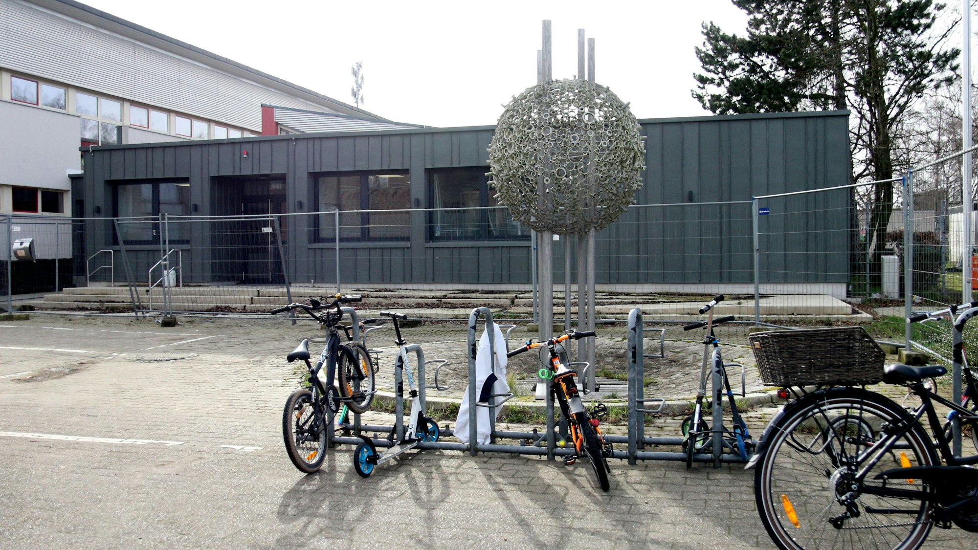 Im Vordergrund ist eine Metallskulptur in Form einer Kugel zu sehen, im Hintergrund der Verwaltungstrakt.