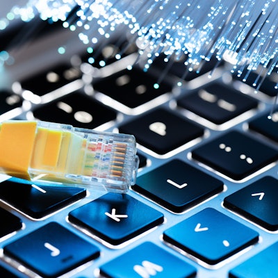 Ein LAN-Kabel liegt auf einer Laptop-Tastatur, im Hintergrund sind Glasfaserkabel zu sehen.