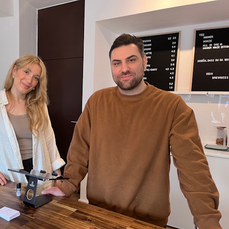 Eine Frau mit blondem Haar und ein Mann mit kurzen dunklen Haaren stehen hinter der Theke in einem Café.